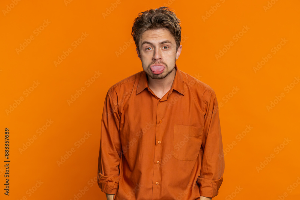 Fototapeta premium Funny Caucasian man showing tongue making faces at camera, fooling around, joking, aping with silly face, teasing, bullying, abuse disrespect. Handsome young guy isolated on orange background indoors