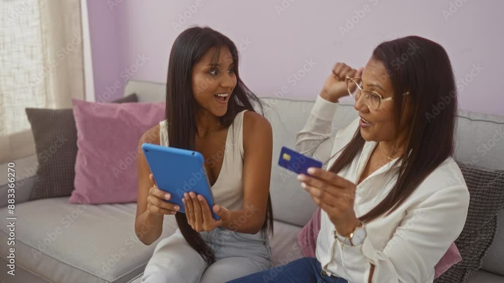 Woman and daughter shopping online at home with a tablet and credit card sitting together on a sofa showing family love and bonding in a cozy living room with pillows. Woman and daughter shopping online at home with a tablet and credit card sitting together on a sofa showing family love and bonding in a cozy living room with pillows.