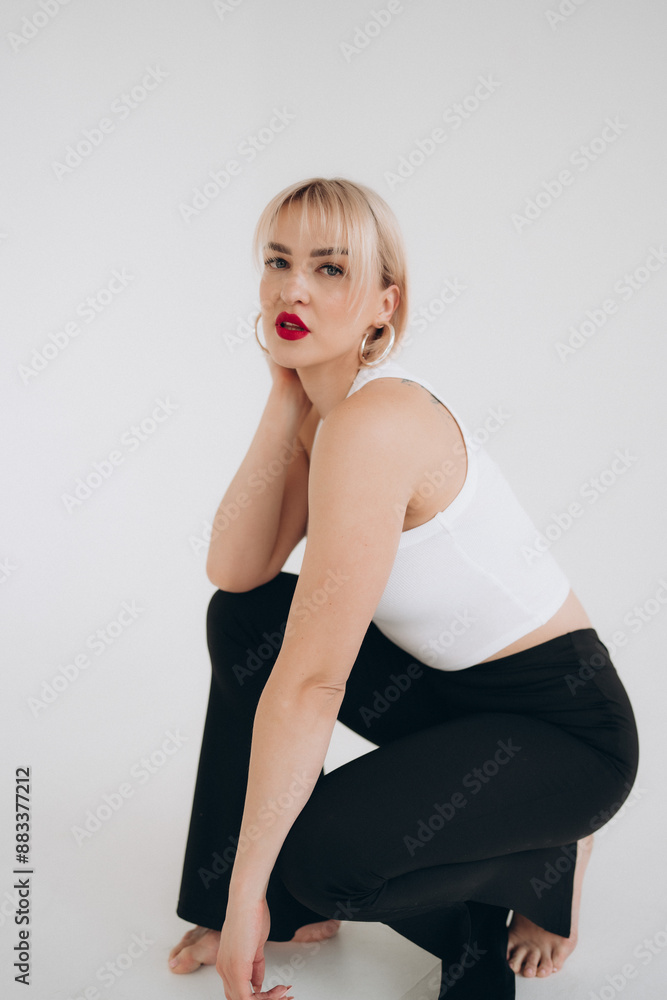 A blonde woman in a white crop top and black pants kneels on the floor, gazing to the side, set against a plain white background. A blonde woman in a white crop top and black pants kneels on the floor, gazing to the side, set against a plain white background.
