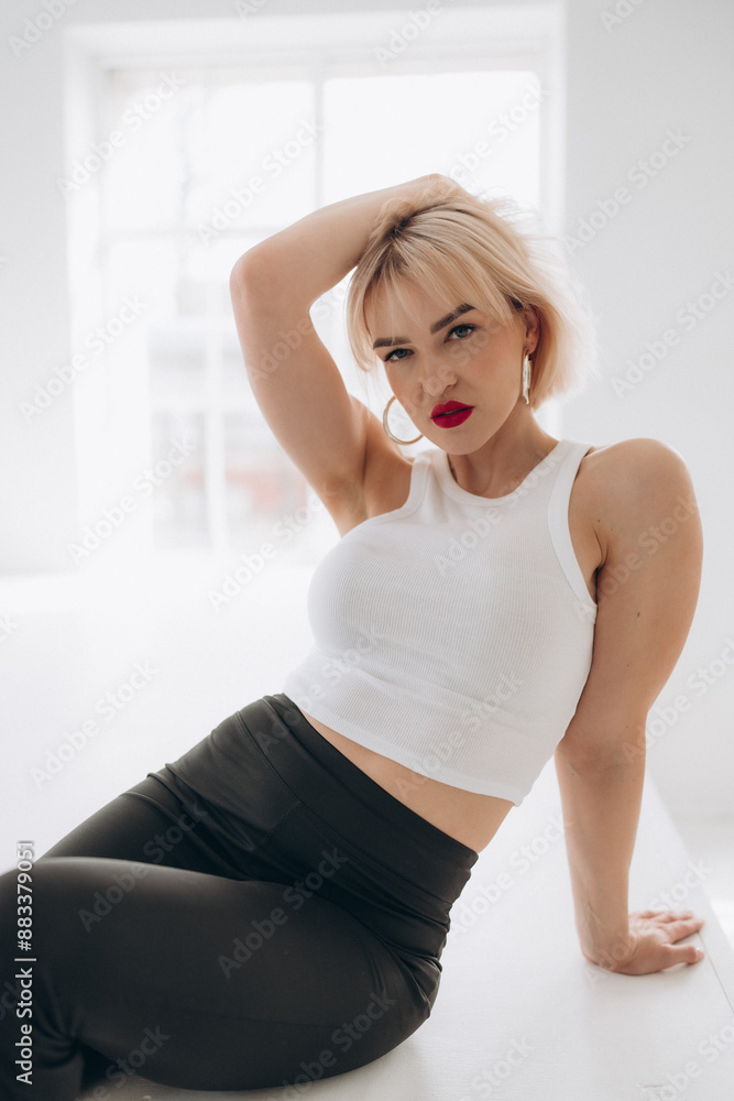 A blonde woman with red lipstick in a white crop top and black pants poses on white steps in a bright, minimalist room. She leans back gracefully. A blonde woman with red lipstick in a white crop top and black pants poses on white steps in a bright, minimalist room. She leans back gracefully.