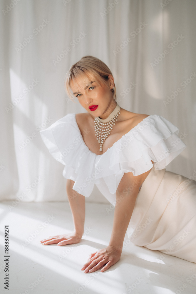 Elegant woman with blonde bob haircut and red lipstick posing on all fours in a white ruffled dress and pearl necklace, against a white background. Copy space... Elegant woman with blonde bob haircut and red lipstick posing on all fours in a white ruffled dress and pearl necklace, against a white background. Copy space...