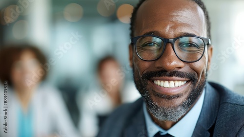 A professional man wearing glasses is smiling warmly in an office setting, which appears bright and modern with other blurred people in the background.