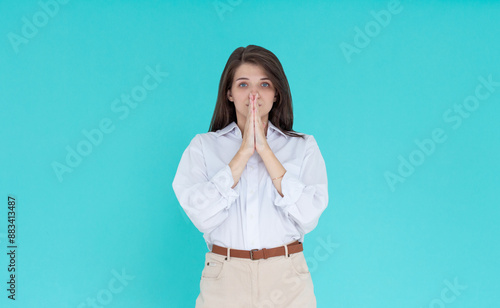 Portrait of imploring girl tilts head, purses lips, looks sadly at camera, makes praying gesture, asks for help and support, begs boyfriend for apologize, wears white shirt, isolated on blue wall