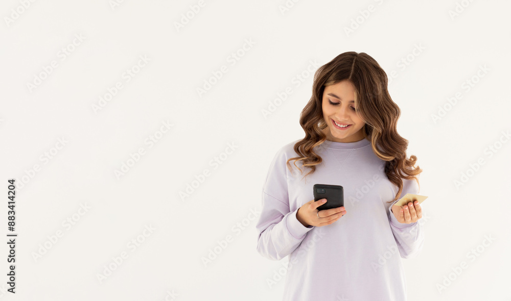 Cheerful young girl showing plastic credit card while using mobile phone isolated over white background, wearing purple sweatshirt Cheerful young girl showing plastic credit card while using mobile phone isolated over white background, wearing purple sweatshirt