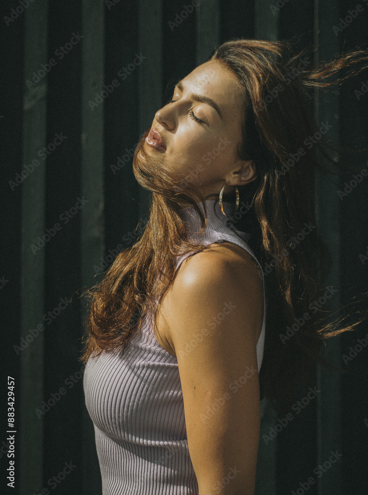 A woman with long hair and hoop earrings poses outdoors with her eyes closed, basking in sunlight, against a dark textured background. A woman with long hair and hoop earrings poses outdoors with her eyes closed, basking in sunlight, against a dark textured background.