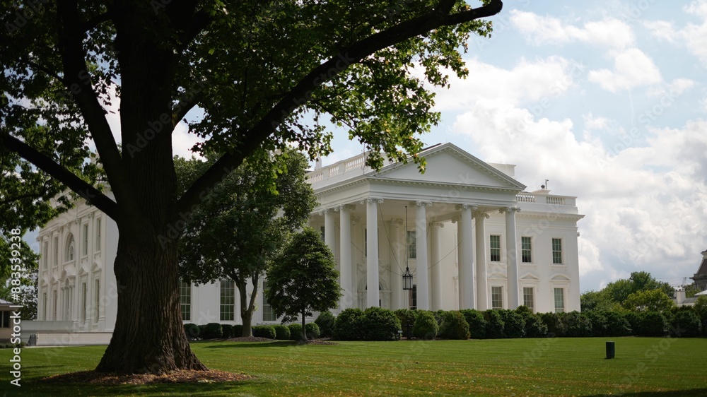 Obraz premium Angled view of the White House with green grass, trees cloudy blue sky in spring.