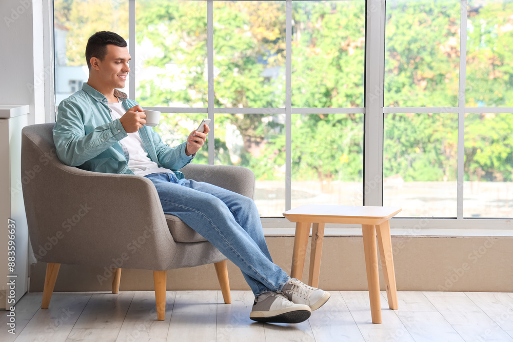 Handsome young man drinking coffee while using mobile phone and sitting on armchair near window