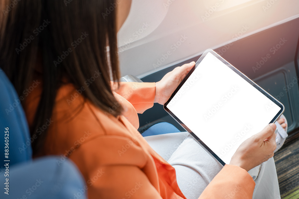 A woman in an orange jacket using a digital tablet with a blank screen while seated on an airplane, ideal for travel, technology, and business themes. A woman in an orange jacket using a digital tablet with a blank screen while seated on an airplane, ideal for travel, technology, and business themes.