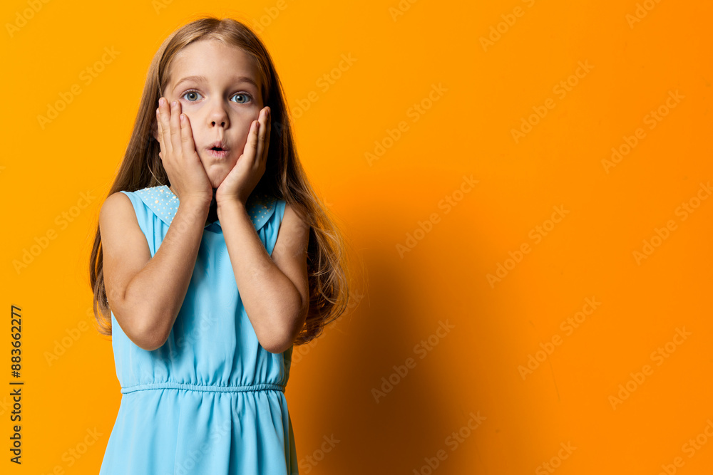 Shy little girl in blue dress hiding her face with hands against vibrant orange background