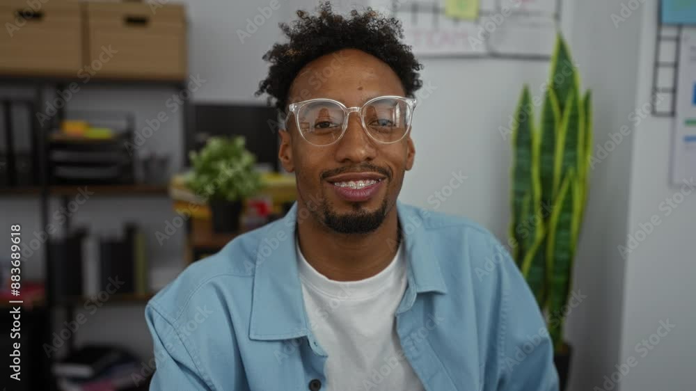 A young black man with glasses is seen in a modern office setting, with indoor plants and office supplies in the background. A young black man with glasses is seen in a modern office setting, with indoor plants and office supplies in the background.