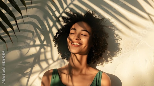 Cheerful young attractive woman in stylish dark-green top enjoys sunny weather, poses near white wall with palm leaf shadow and smiles.  photo