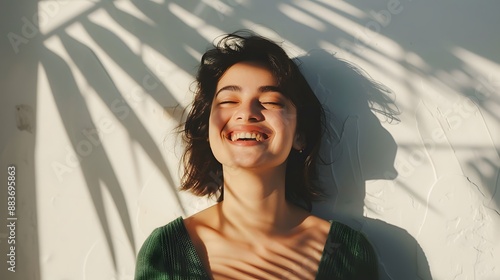 Cheerful young attractive woman in stylish dark-green top enjoys sunny weather, poses near white wall with palm leaf shadow and smiles.  photo