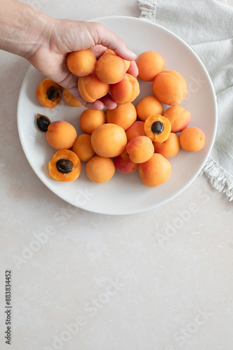Top down close view of two hands of a woman, holding halved apricot over a plate with fresh ripen apricots, with copy space