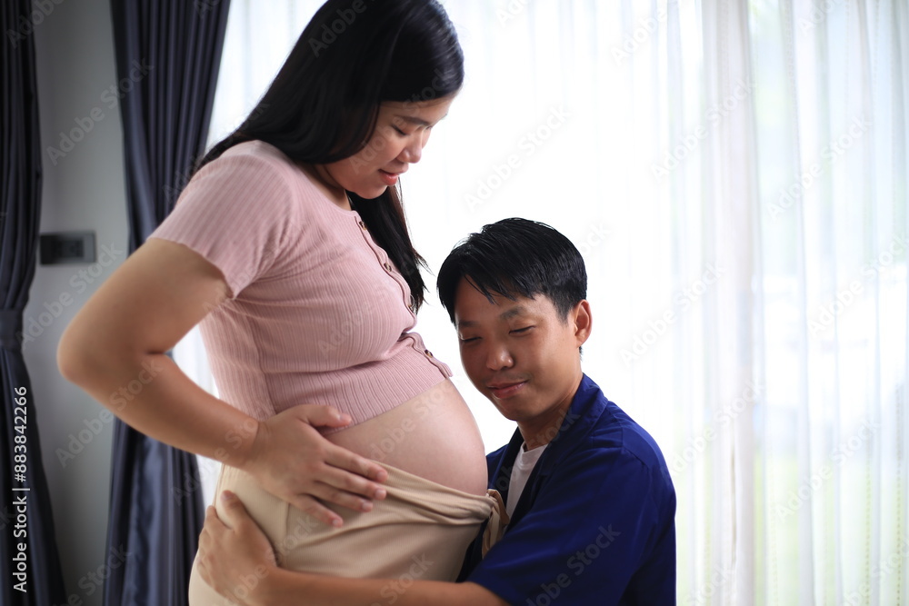 Fototapeta premium An asian couple seems very happy. A handsome husband is embracing his beautiful pregnant wife next to the window in the living room.Pregnant woman with husband holding ultrasound baby image.