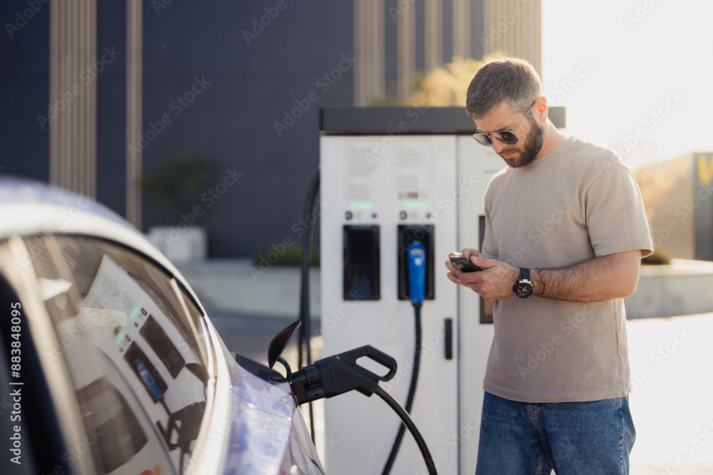 A middle-aged man checks his smartphone while his electric car charges at a public charging station during a sunny day. A middle-aged man checks his smartphone while his electric car charges at a public charging station during a sunny day.