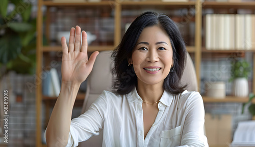 Beautiful Middle-Aged Asian Woman Sitting at Desk in Home Office Waving Hello on Video Call with Family or Friends, Smiling at Camera