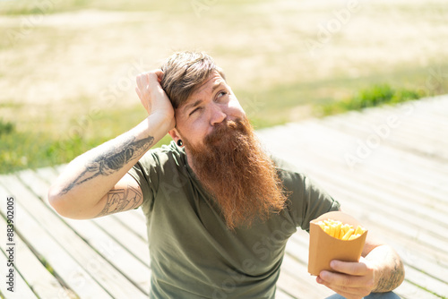 Redhead man with beard holding fried chips at outdoors having doubts and with confuse face expression photo