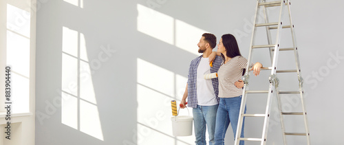 Family couple doing renovations at home. Young man and woman change color of walls in living room. Banner with happy husband and wife standing by ladder and looking at freshly painted light gray wall photo