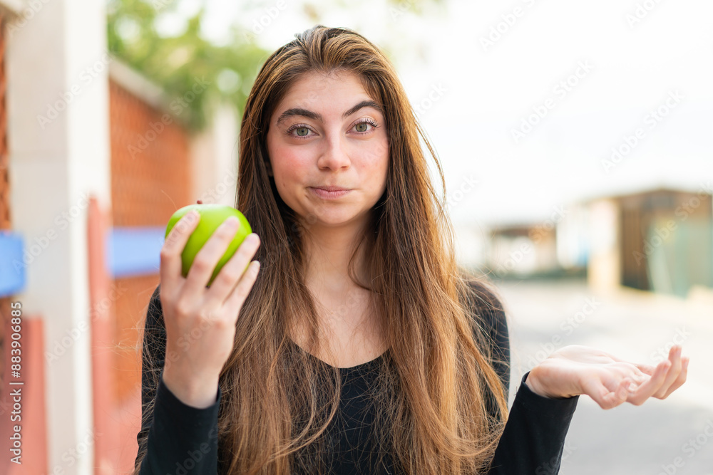 Young pretty caucasian woman with an apple at outdoors making doubts gesture while lifting the shoulders Young pretty caucasian woman with an apple at outdoors making doubts gesture while lifting the shoulders