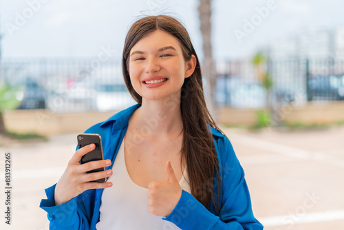 Young pretty Ukrainian woman using mobile phone at outdoors with thumbs up because something good has happened Free Download Young pretty Ukrainian woman using mobile phone at outdoors with thumbs up because something good has happened - GFXLoad