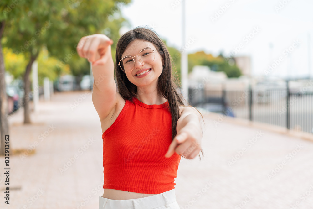 Young woman with glasses at outdoors points finger at you while smiling Young woman with glasses at outdoors points finger at you while smiling