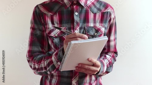 man with checkered shirt takes notes, he's against a white background
