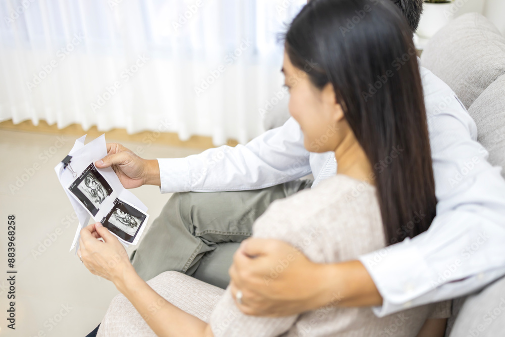A woman hold ultrasound image with husband in a home setting. Pregnancy with baby ultrasound after successful infertility technology with ivf technique. Asian couple in living room looking at baby.