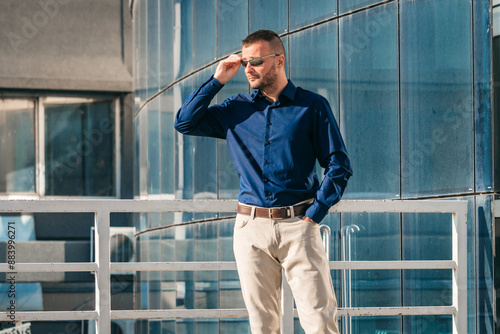 Man in Blue Shirt and Sunglasses Stands on a Balcony With a City View