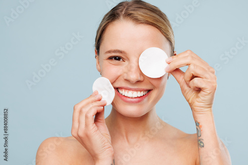 Beautiful smiling half naked topless young woman with nude make up clean face cover close eye with cotton pad isolated on plain blue color background. Skin care healthcare cosmetic procedures concept. photo