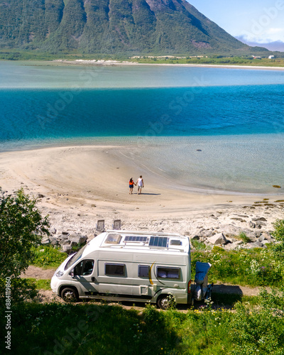 A white camper van is parked near a sandy beach in Norway, with a couple walking on the shore in front of a mountain backdrop and turquoise water. men and women at the Lofoten Norway