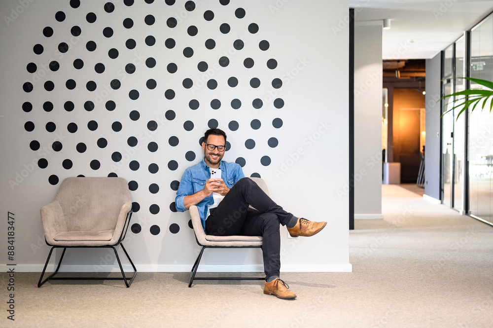Full length of happy young man messaging over smart phone while sitting comfortably on office chair Full length of happy young man messaging over smart phone while sitting comfortably on office chair