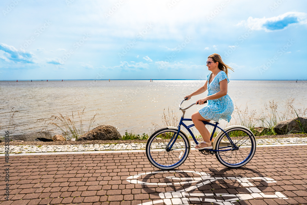Fototapeta premium Active summer holidays. Woman with long hair wearing blue dress riding blue cruiser bike on bicycle path on seafront on sunny day. Side view