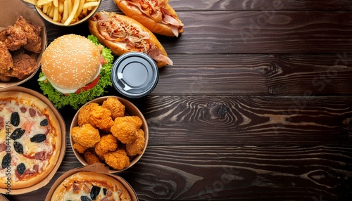 Top view of various takeout or delivery foods like hamburgers, pizza, fried chicken, and sides on a dark wooden background with ample copy space
