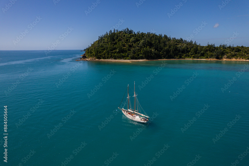 Aerial view of a sailboat anchored near a lush, green island with turquoise waters under a clear blue sky, capturing the essence of a tranquil tropical paradise.
