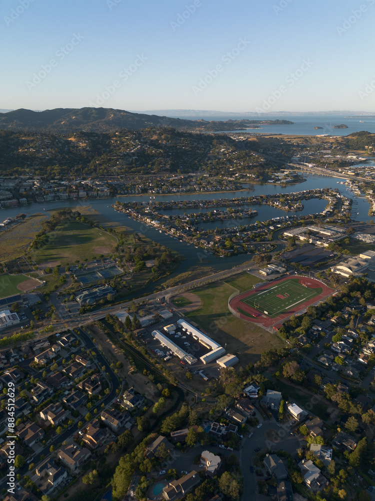 Fototapeta premium Arial wide of town with river running through along bay, lots of blue water and green trees, golden hour sunset shining