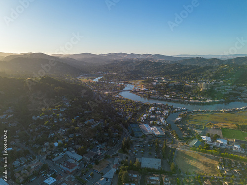 Arial wide of town with river running through, lots of blue water and green trees, golden hour sunset shining