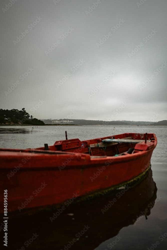 Fototapeta premium Red wooden boat floating on a calm lake under an overcast sky