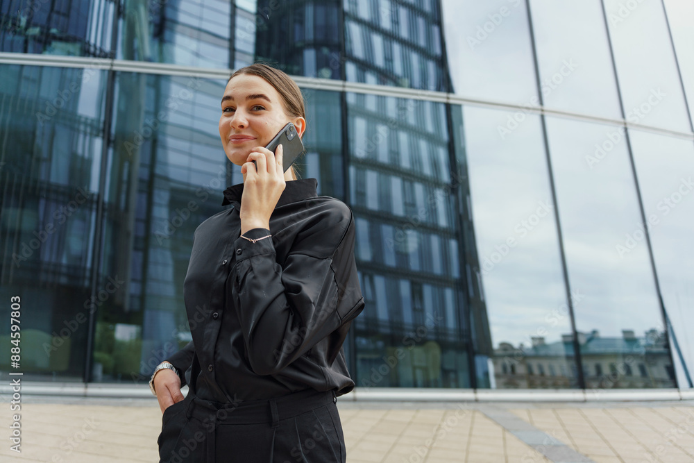 A confident businesswoman in a black outfit talks on her phone, with modern glass architecture behind her. A confident businesswoman in a black outfit talks on her phone, with modern glass architecture behind her.