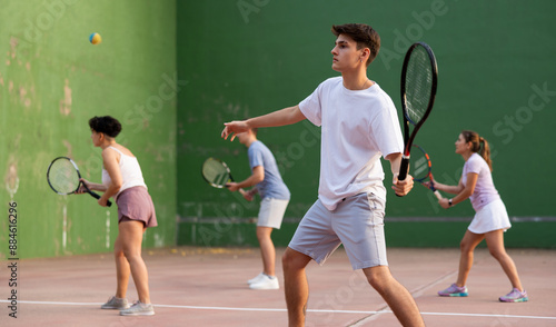 Athletic young hispanic guy playing frontenis on open court on summer day, hitting ball with strung tennis racquet to score to opposing team. Popular sports photo