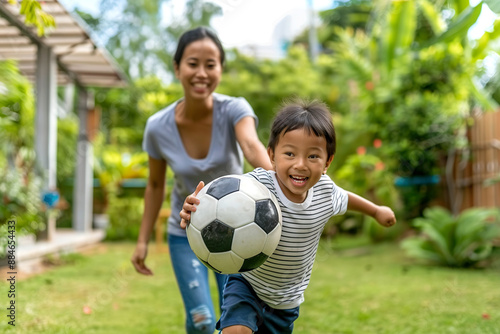 A young boy smiles as he runs with a soccer ball in a green grassy backyard. An adult woman walks behind him, watching him.