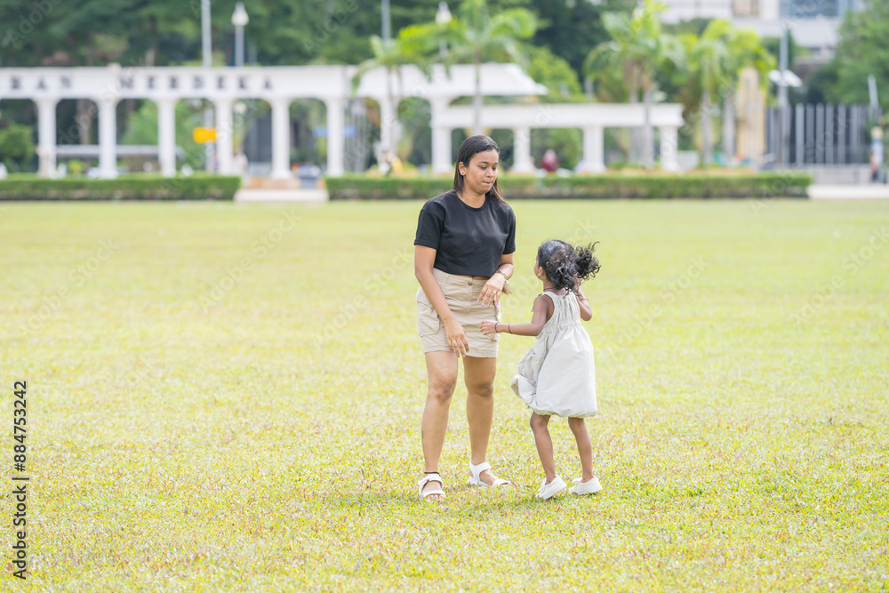 A thirty-something Indian Malaysian mother joyfully playing on the expansive lawn at Merdeka Square in Kuala Lumpur, Malaysia, while holding and running around with her five-year-old daughter. A thirty-something Indian Malaysian mother joyfully playing on the expansive lawn at Merdeka Square in Kuala Lumpur, Malaysia, while holding and running around with her five-year-old daughter.