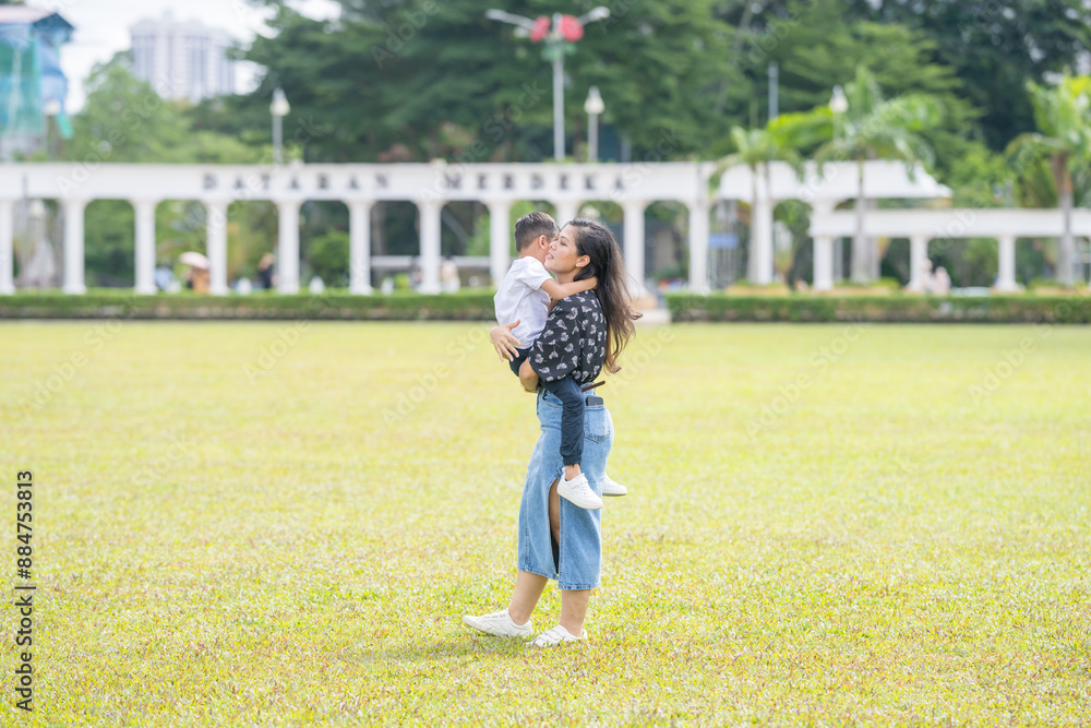 A 30-something Indian Malaysian mother joyfully playing on the expansive lawn at Merdeka Square in Kuala Lumpur, Malaysia, while holding her 7-year-old son. A 30-something Indian Malaysian mother joyfully playing on the expansive lawn at Merdeka Square in Kuala Lumpur, Malaysia, while holding her 7-year-old son.