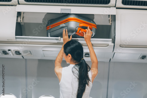 Obraz na płótnie Young Female Tourist Storing Luggage in Overhead Compartment on Airplane During