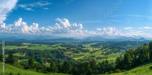 panoramic view of the Bavarian Alps with green meadows and villages, clouds in the sky, mountains far in the distance, trees all around, summer time, blue skies, photo-realistic, taken on a Canon EOS 