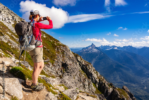 Adventurous athletic female hiker taking a photo with her cellphone of jagged mountains in the in the Pacific Northwest.
