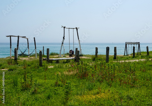 Yangyang-gun, Gangwon-do, South Korea - June 25, 2023: Summer and morning view of grass and a couple sitting on swing against sea at Jeongam Beach 