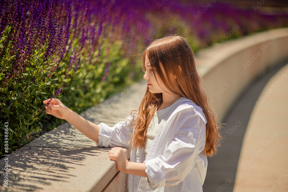 Naklejka premium A young girl is basking in the warm rays of the sun, enjoying the beauty of the park, overgrown with bright purple sage. Her white shirt and blue trousers complete the landscape