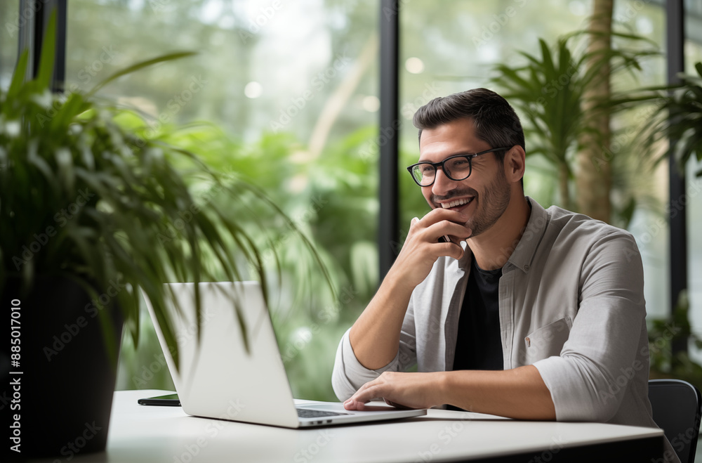 A smiling man with glasses works on his laptop at a modern office with green plants and large windows. A smiling man with glasses works on his laptop at a modern office with green plants and large windows.