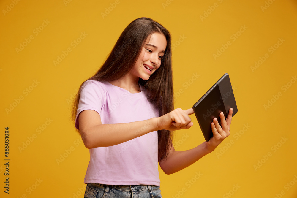 Happy smiling teen girl with brown long hair, in pink t-shirt, using tablet for leisure and education against yellow studio background. Concept of human emotions, childhood, youth, education Happy smiling teen girl with brown long hair, in pink t-shirt, using tablet for leisure and education against yellow studio background. Concept of human emotions, childhood, youth, education