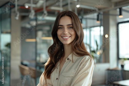 A young, beautiful brunette woman stands in a modern office, smiling happily as she looks directly at the camera, exuding confidence and professionalism.